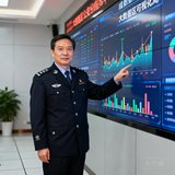 成都某区公安分局指挥中心张总监的半身照片，他身穿警服站在大数据可视化大屏前，手指向屏幕上的实时数据